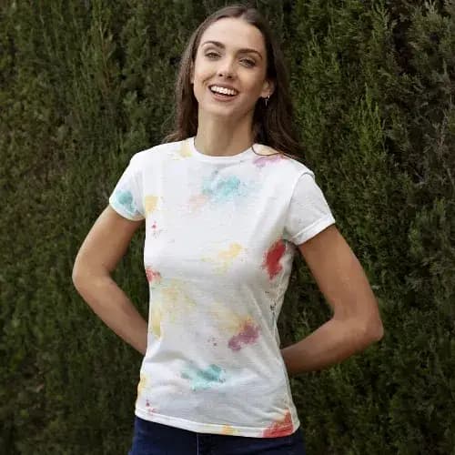 A woman in a white t-shirt featuring a vibrant, multi-coloured splatter design, standing outdoors against greenery. Hearvin - Image 1