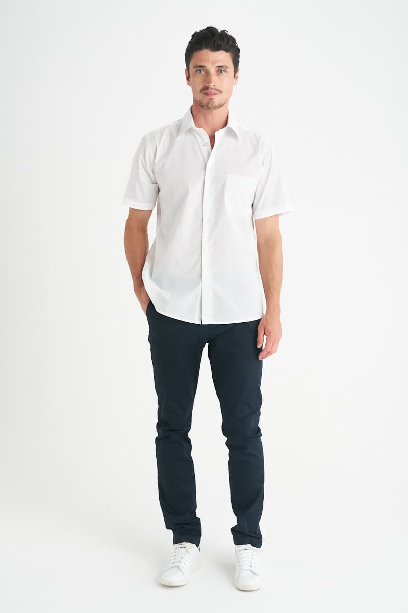 A man wearing a white short sleeve button-up shirt with a chest pocket, paired with dark trousers and white shoes. Rodeo - Mens Short Sleeve Shirt - Image 1
