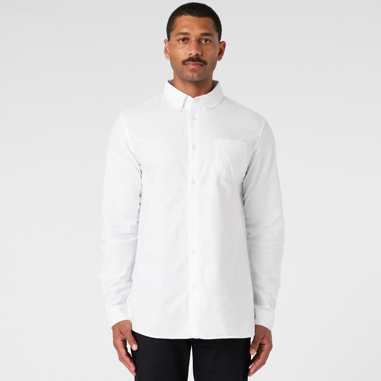 A white button-up Oxford shirt on a model, featuring a chest pocket and long sleeves. The shirt has a logo. Oxford Shirt - Image 1