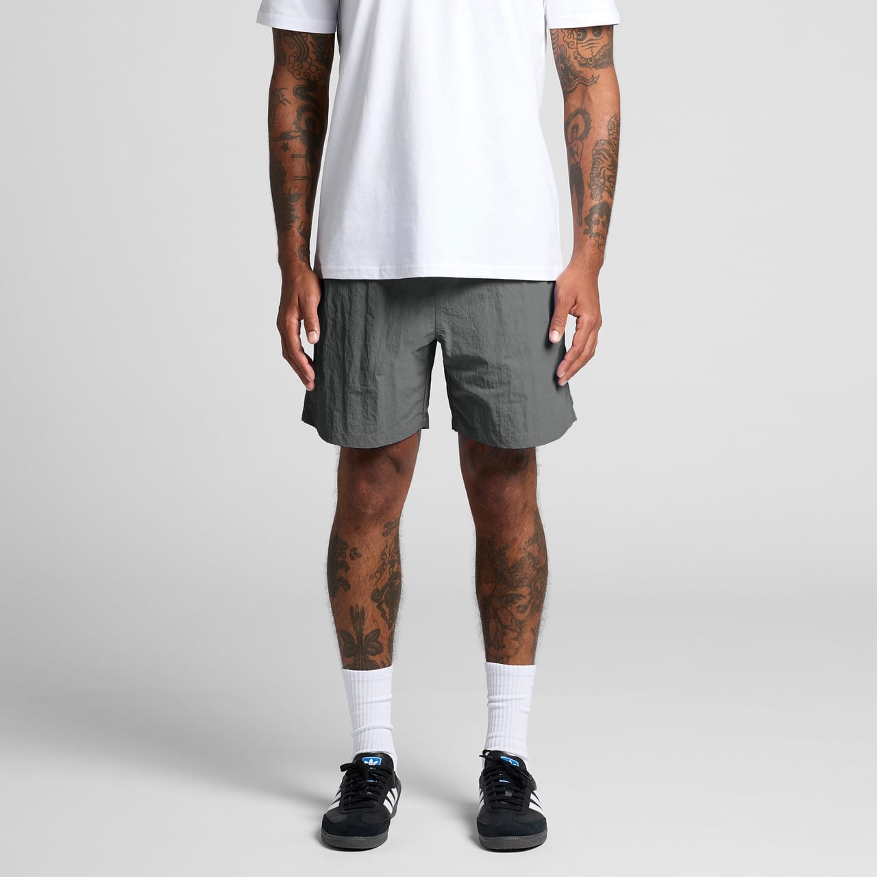 A person stands wearing grey swim shorts, paired with a white t-shirt and black sneakers. The shorts feature a logo. Swim Shorts 17" - Image 1