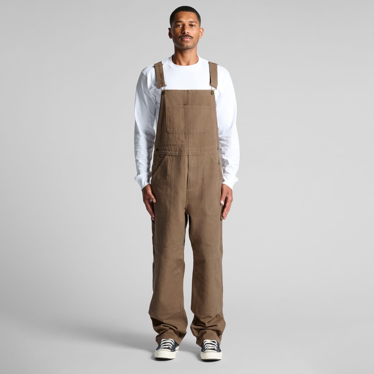 Khaki canvas overalls worn by a model, featuring adjustable straps and a relaxed fit. The model pairs them with a white long-sleeve shirt. Canvas Overalls - Image 1