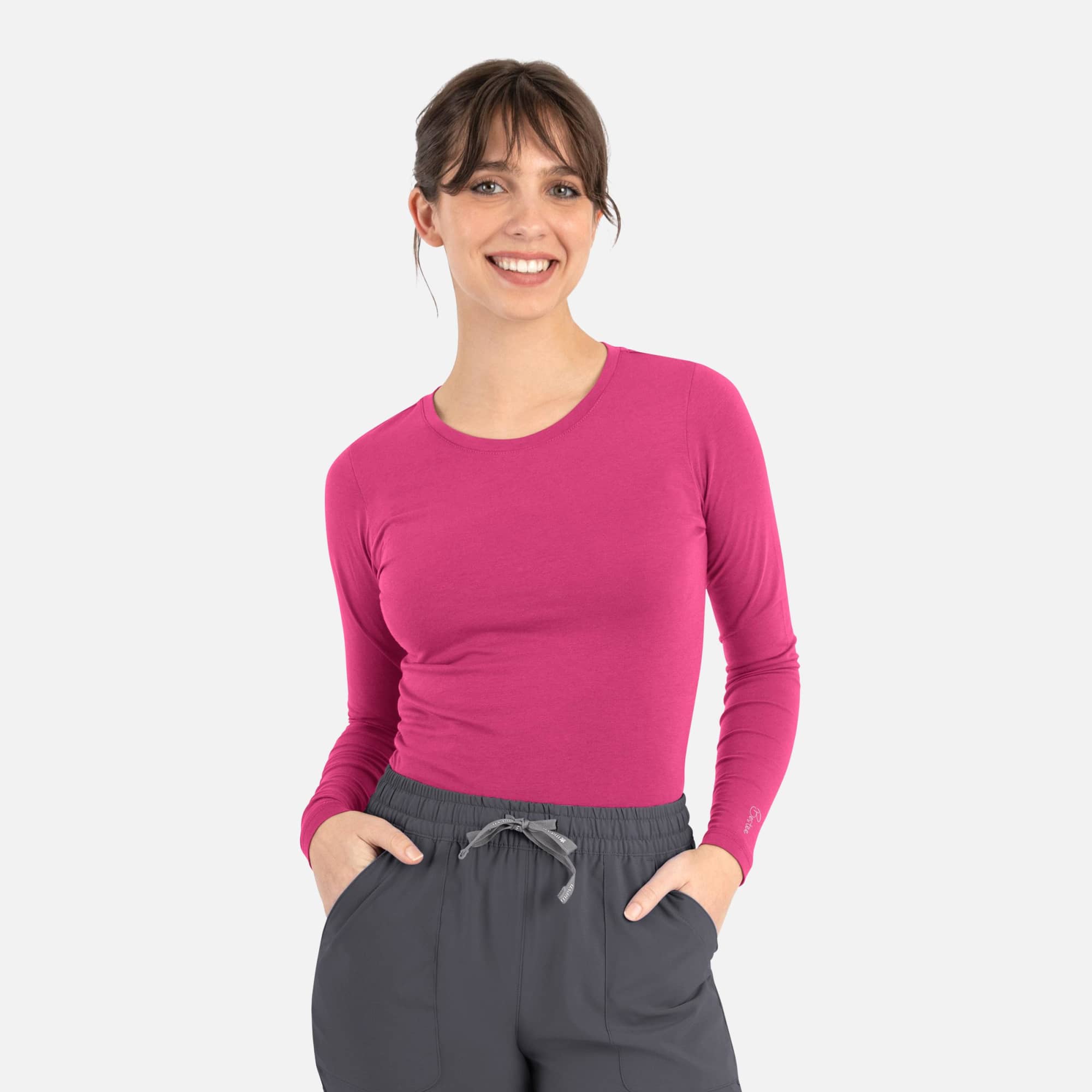 A woman wearing a long-sleeve, bright pink underscrub tee and grey pants, smiling while posing with her hands in her pockets. Bestee Womens Underscrub Tee - Image 1