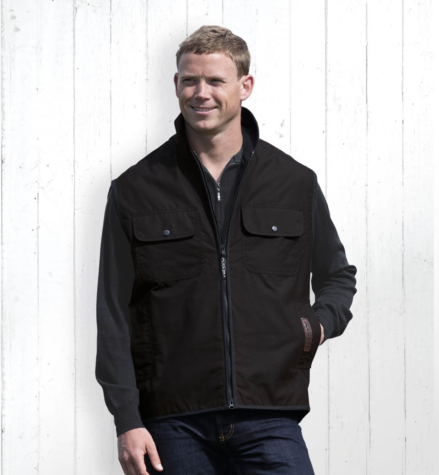 A man wearing a black oilskin vest with two front pockets, standing in front of a white wooden background. Agri Station Ranger Oilskin Vest - Image 1