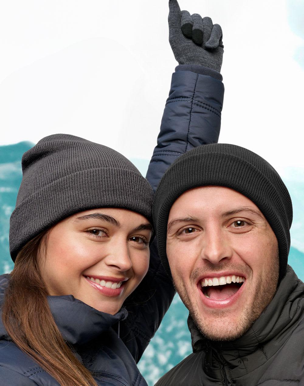 A grey beanie is worn by a man and a woman, both smiling and wearing winter jackets against a snowy backdrop. Roll Up rPET Beanie - Image 1