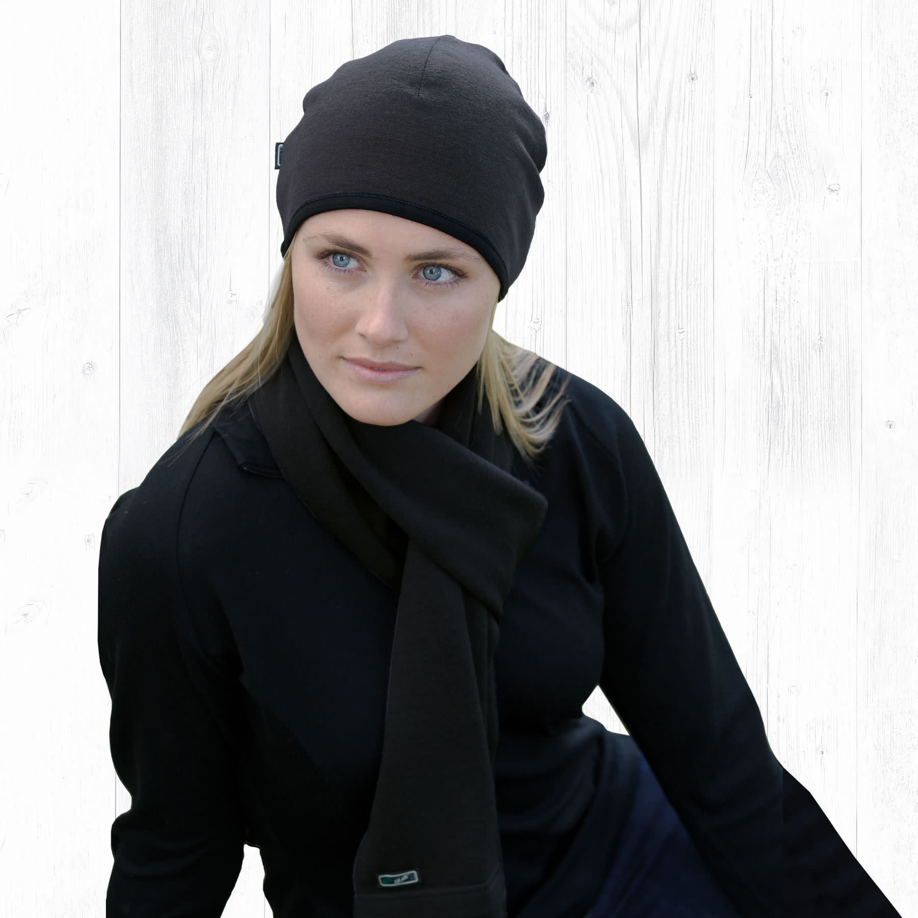 A woman wearing a black Merino Arctic Scarf and matching beanie, seated against a light wooden background. Merino Arctic Scarf - Image 1