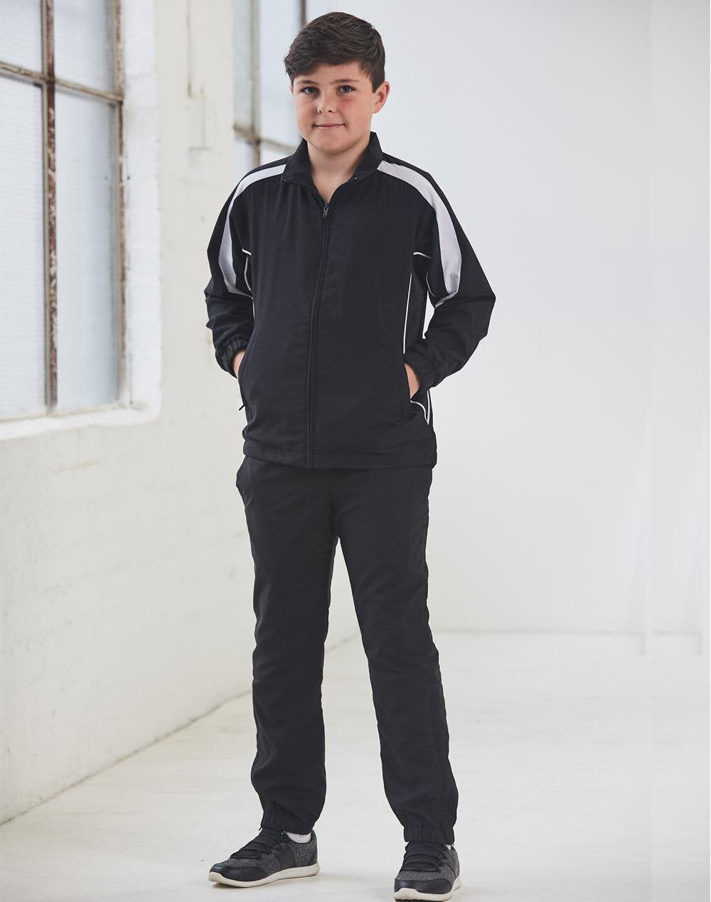 A black track jacket with white accents, featuring a zip front and side pockets, modelled by a young boy. Kids Warm Up Jacket - Image 1