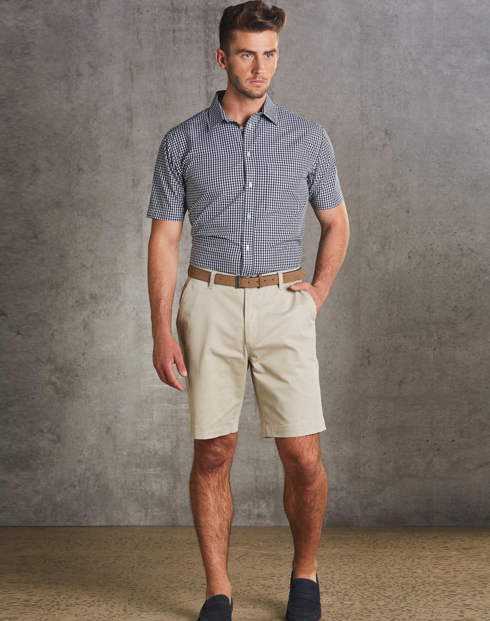 Men's chino shorts in beige, paired with a checked short-sleeve shirt and a brown belt. The model stands against a textured grey backdrop. Men's Chino Shorts - Image 1