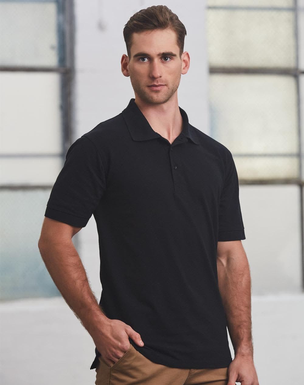 A man in a black polo shirt stands against an industrial background, featuring short sleeves and a standard collar. mens S/S pique polo - Image 1
