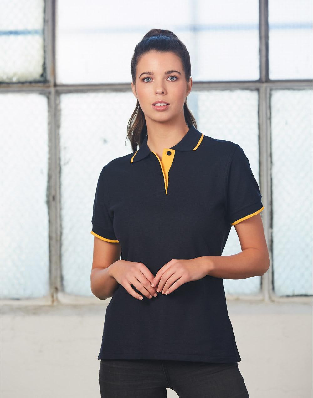 A black polo shirt with yellow accents, featuring a collar and a logo. The model stands in front of a window. Ladies' Poly/Cotton Contrast Pique S/S Polo - Image 1
