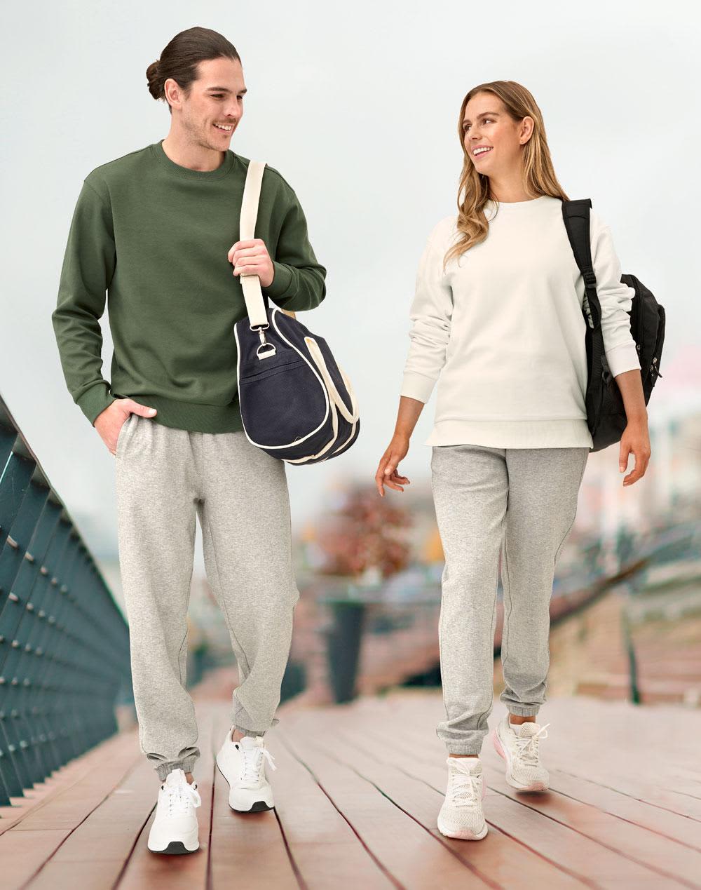 A man in a green sweatshirt and grey sweatpants walks alongside a woman in a white sweatshirt and grey sweatpants, both carrying bags. Unisex Airlayered CVC Sweatpants - Image 1