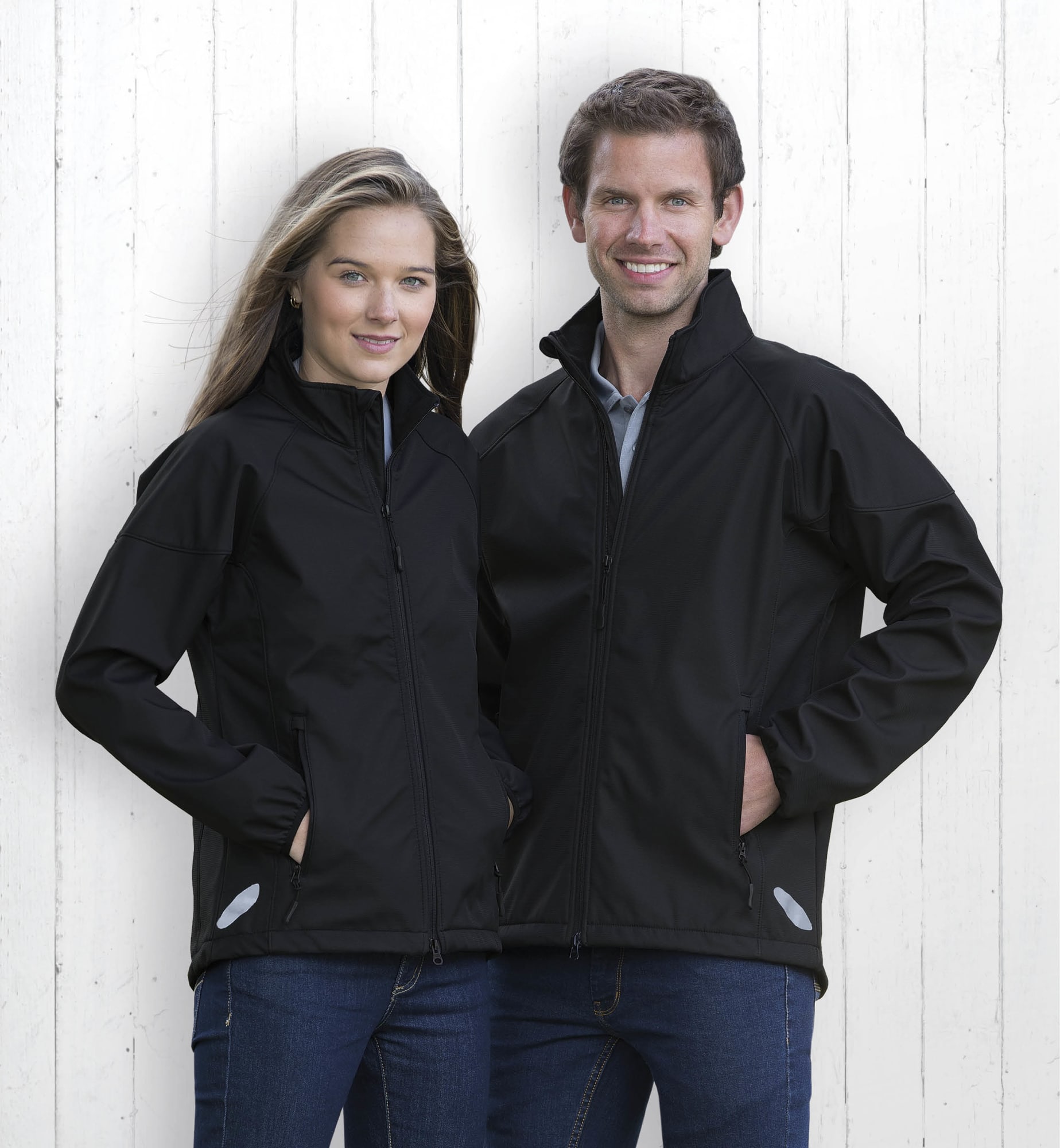 A couple stands in front of a white wooden background, wearing black soft shell jackets. The jackets feature a logo. Traverse Jacket - Image 1