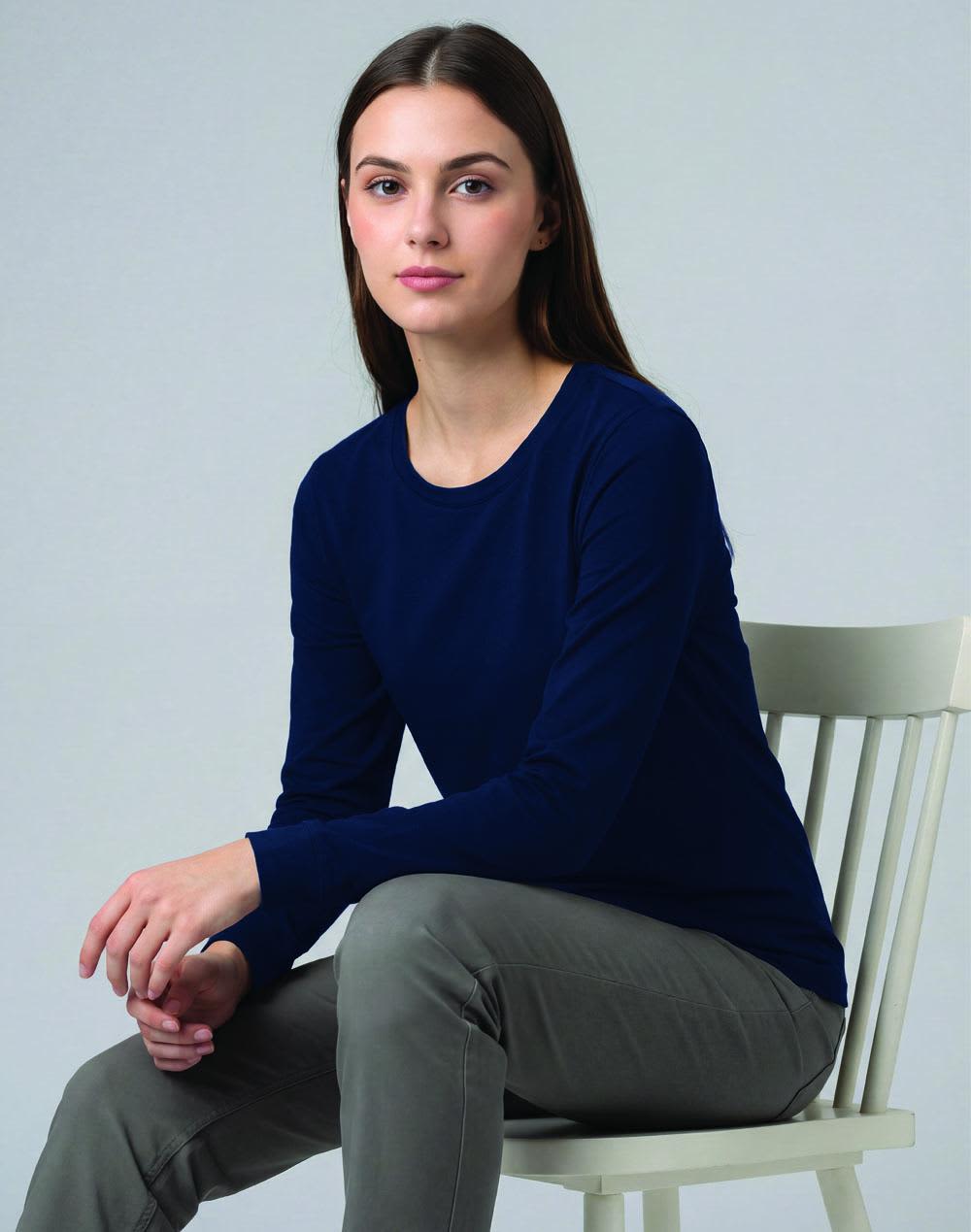 A woman wearing a long-sleeve grey tee shirt and a red and black backpack stands outdoors against a forest backdrop. Ladies' Premium Cotton Face L/S Tee Shirt - Image 1
