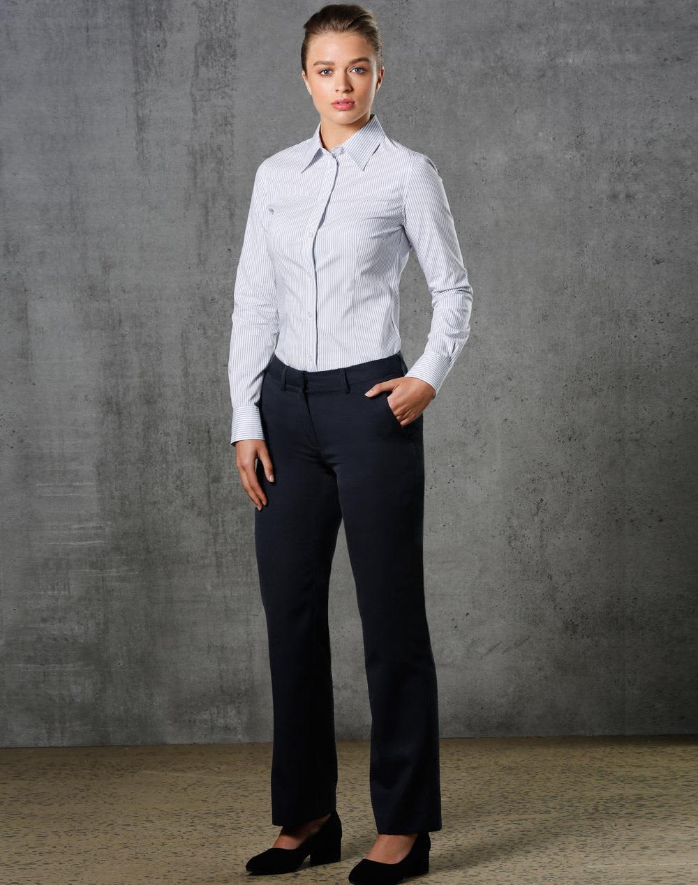 A model wearing ladies' permanent press pants in black, paired with a light blue button-up shirt, standing against a textured backdrop. Ladies Permanent Press Pants - Image 1