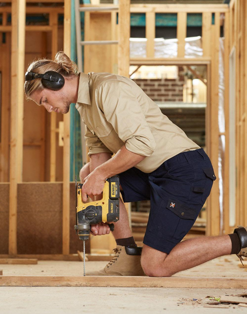 A man in khaki and navy work shorts operating a drill, wearing ear protection, in a construction setting. AIWX Workwear Short - Image 1