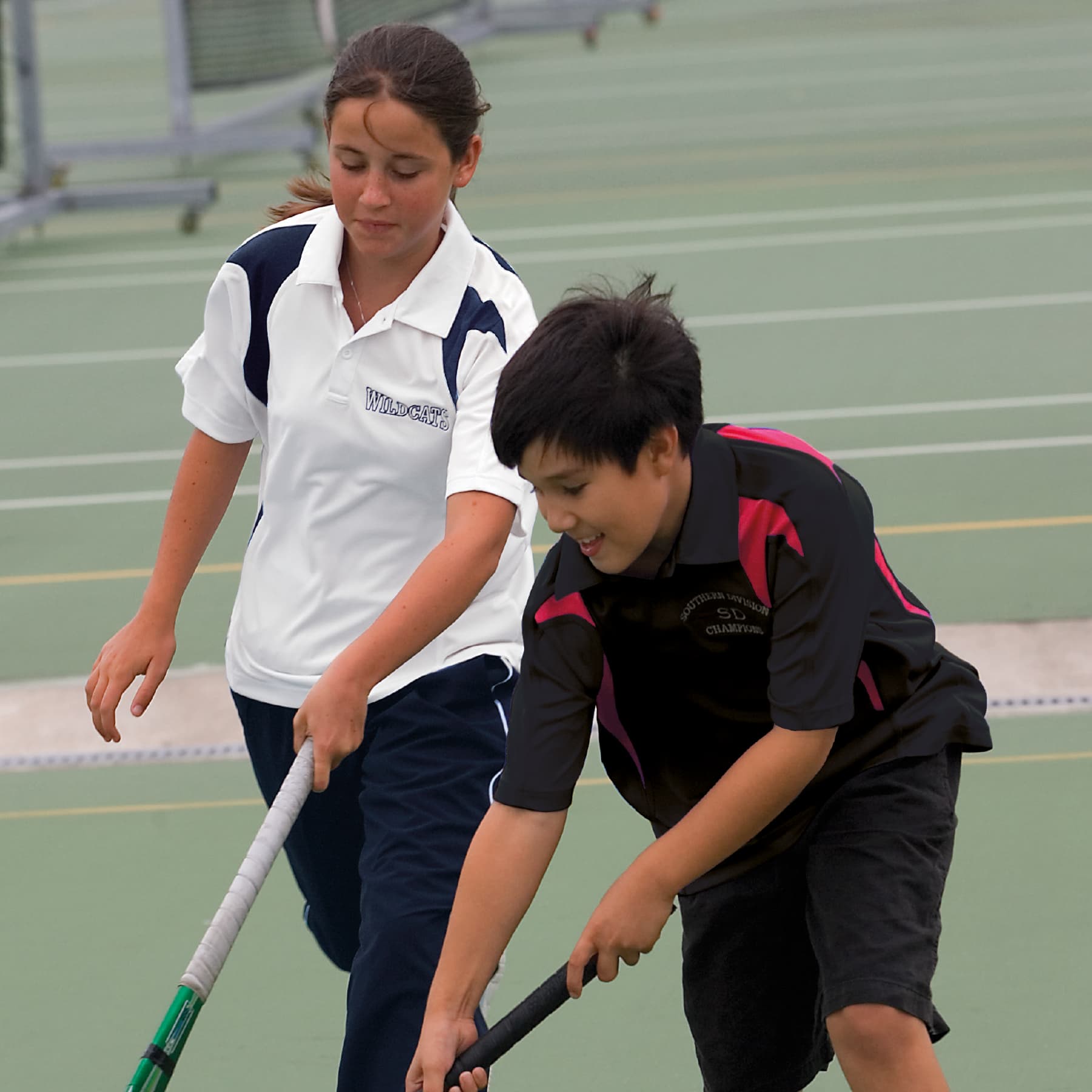 A white polo shirt with navy accents and a dark polo shirt with red accents, worn by two young athletes on a tennis court. Youth Dri Gear Spliced Zenith Polo - Image 1