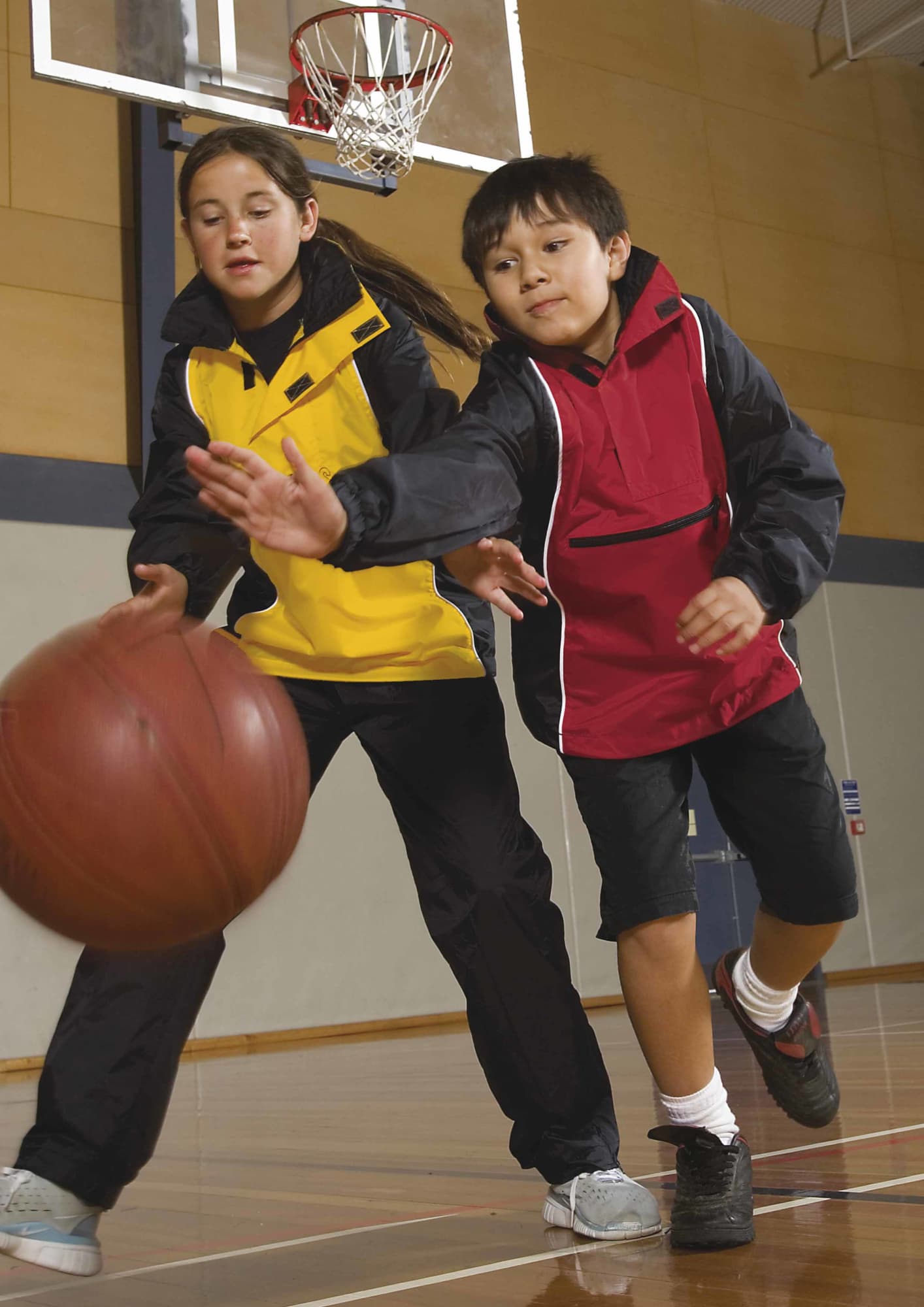 A boy in a red rain jacket and a girl in a yellow rain jacket play basketball indoors. Both jackets have branding. Youth Nylon Jac Pac - Image 1