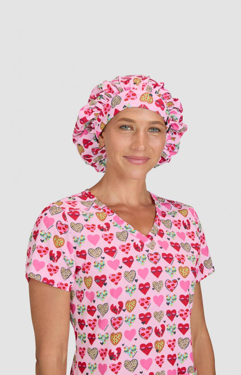 A pink bouffant cap with a heart print, worn by a model in a matching scrubs top with a heart design. Bouffant Cap (Print) - Image 1