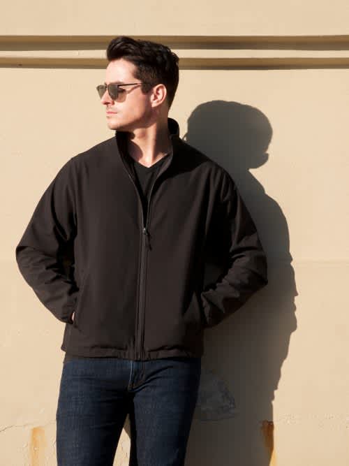 A man wearing a black soft shell jacket stands against a wall, with sunglasses and a shadow. Alta - Mens - Image 1