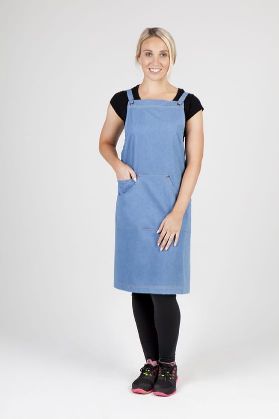 A woman wears a full body denim apron in blue, featuring straps and a front pocket, with black leggings and shoes. Full Body Cotton/Denim Apron - Image 1