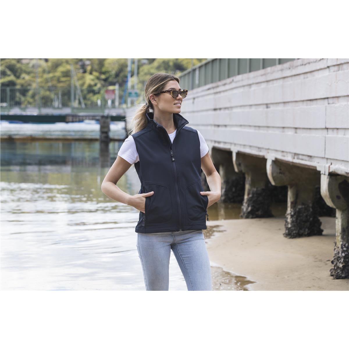 A woman wearing a black softshell vest over a white T-shirt, standing near the water with hands in pockets. Morgan Women's Softshell Vest - Image 1