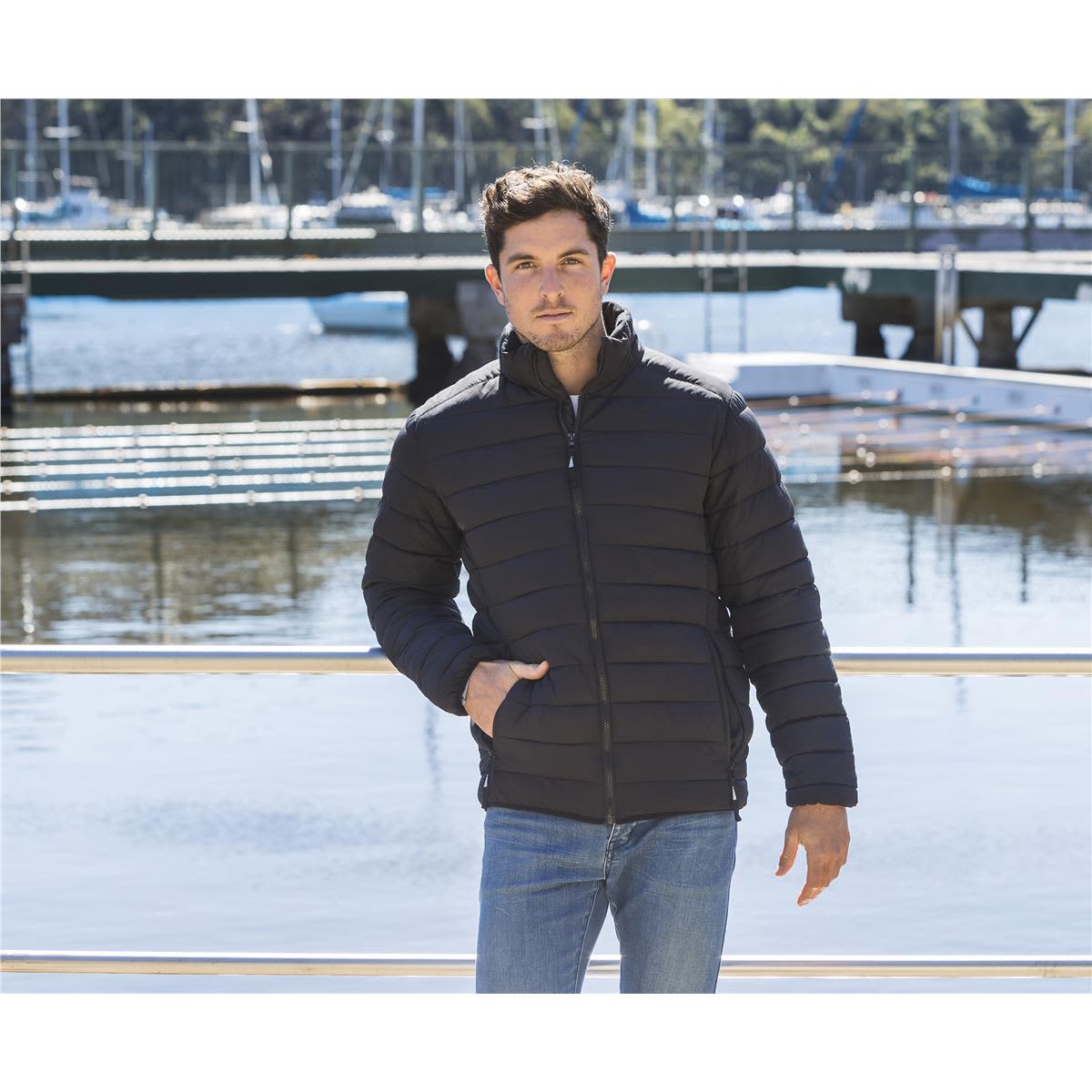 A unisex black puffer jacket with a zip front and side pockets, modelled by a man beside a waterfront. Hudson Unisex Puffer Jacket - Image 1
