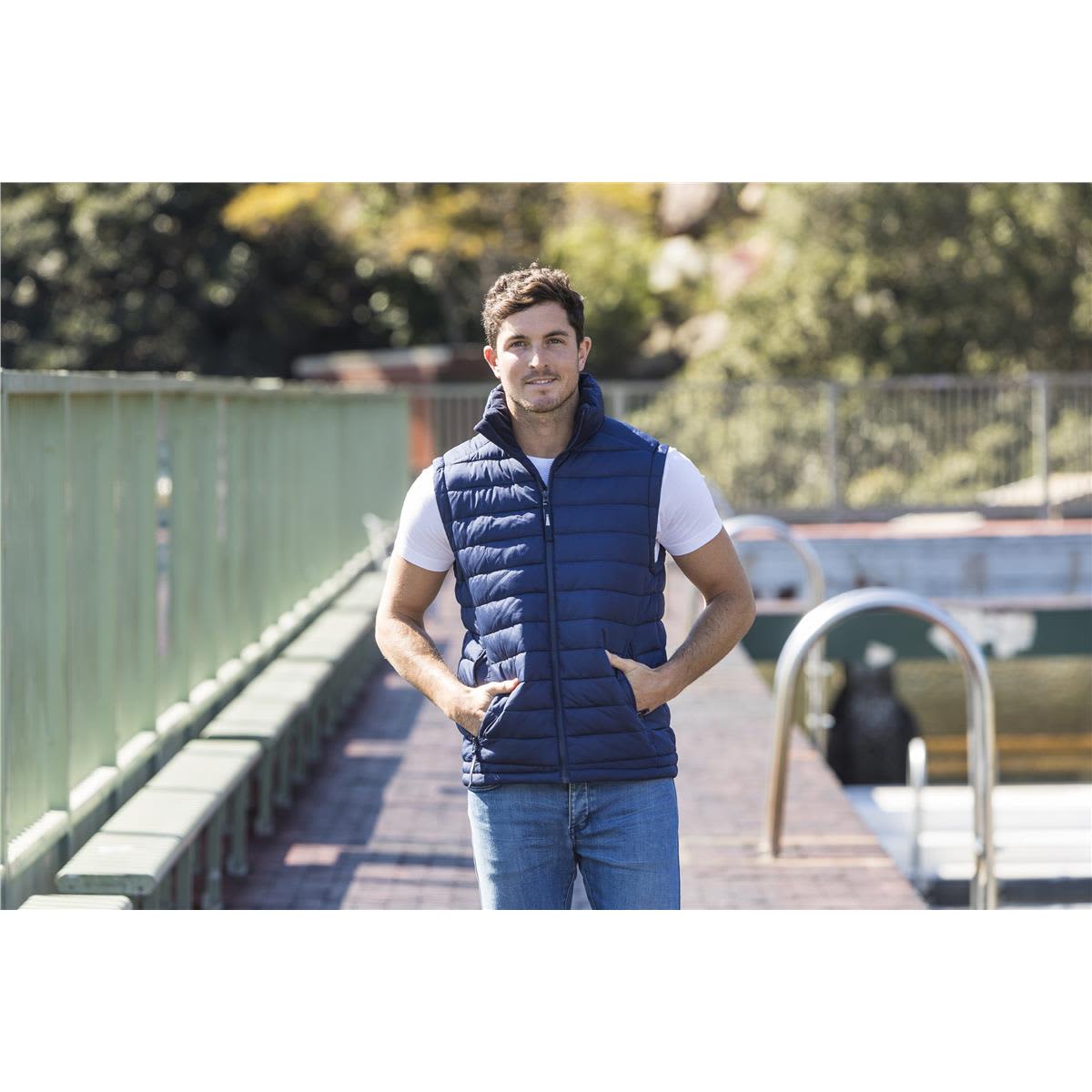 A man stands by a waterway wearing a navy blue unisex puffer vest over a white t-shirt and jeans. Loma Unisex Puffer Vest - Image 1