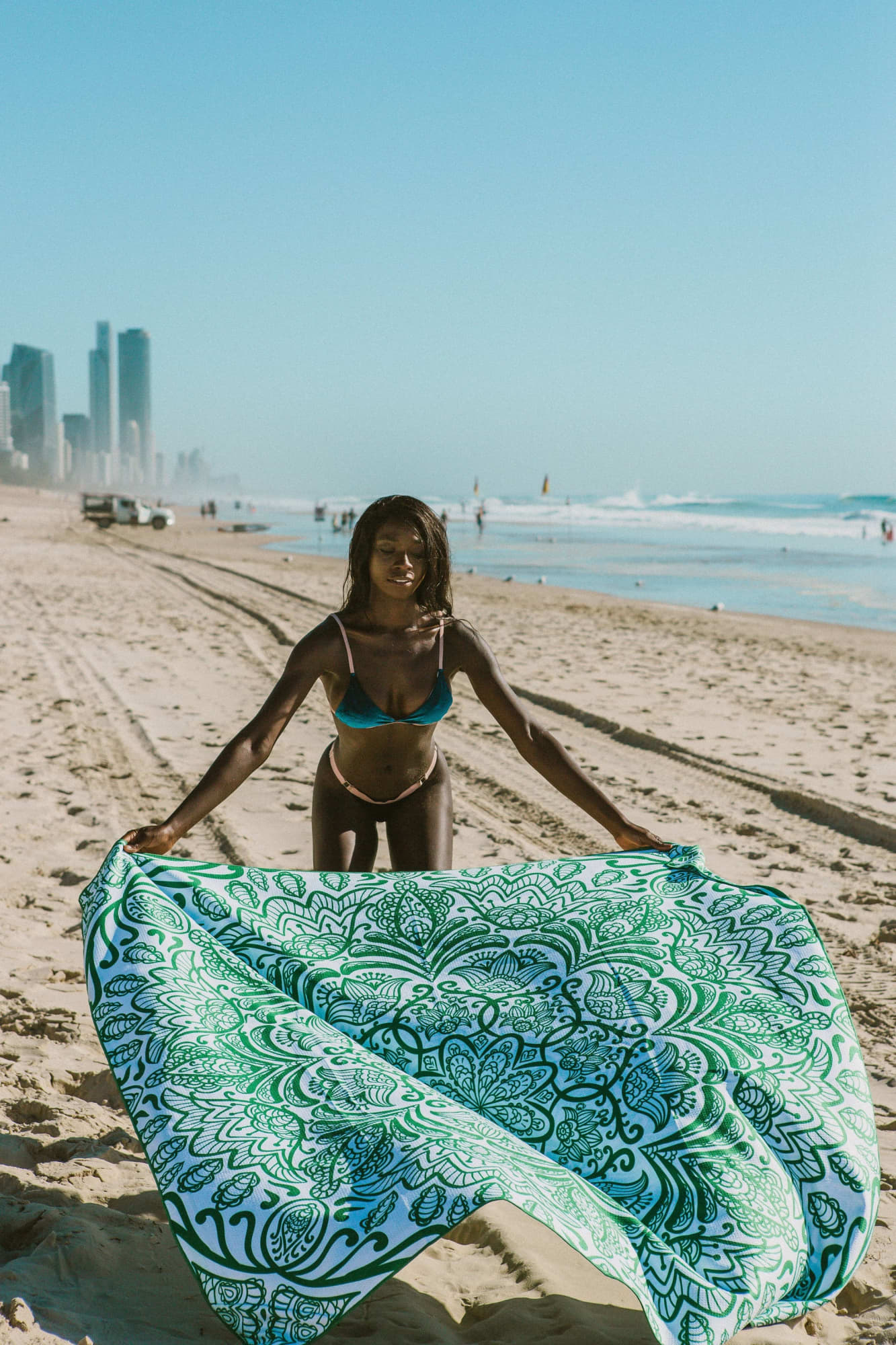 A woman holds a large green and white patterned towel at the beach, with city buildings in the background. Double Size rPET Sand Free Towel - Image 1