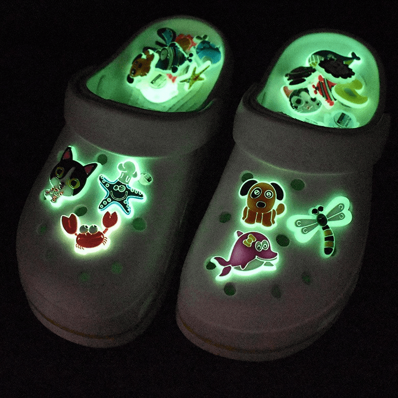 Glow-in-the-dark Croc charms featuring various colourful designs, set against white clogs in a dark background. Personality Glow in Dark Croc Charms - Image 1