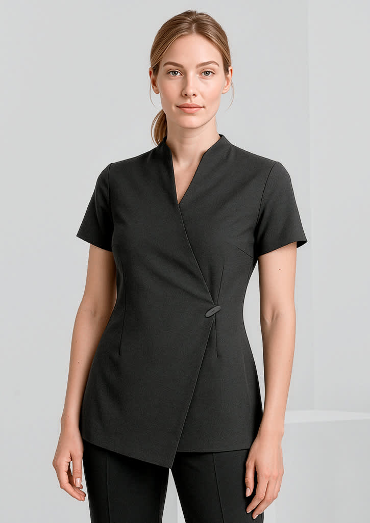 A woman is wearing a black spa tunic with a wrap design and short sleeves. The tunic features a simple button detail. Womens Spa Tunic - Image 1