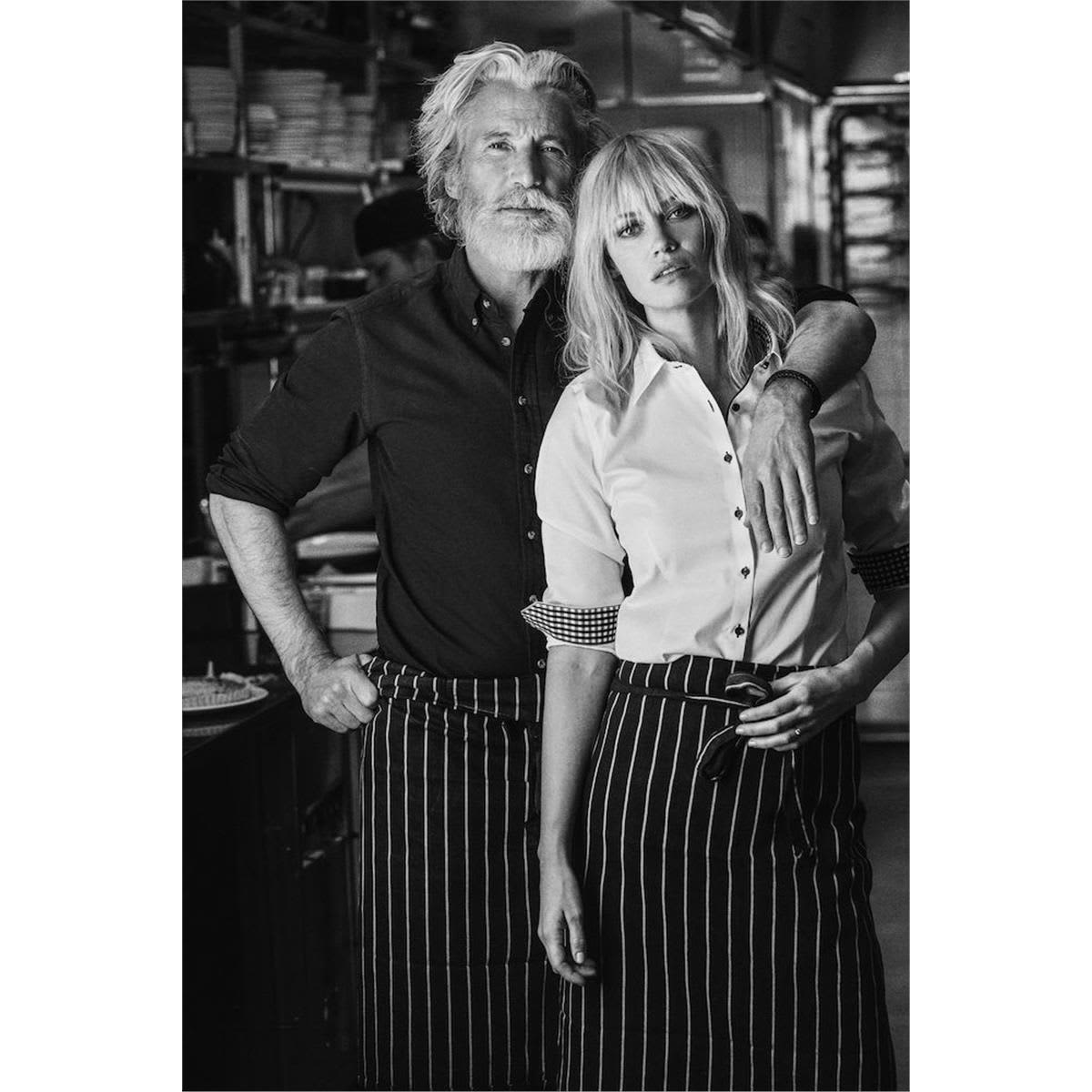 A man and a woman pose together in a kitchen setting, both wearing stylish button-up shirts. The woman’s shirt is white, and the man's is dark. Red Bow 20 Women's Shirt - Image 1