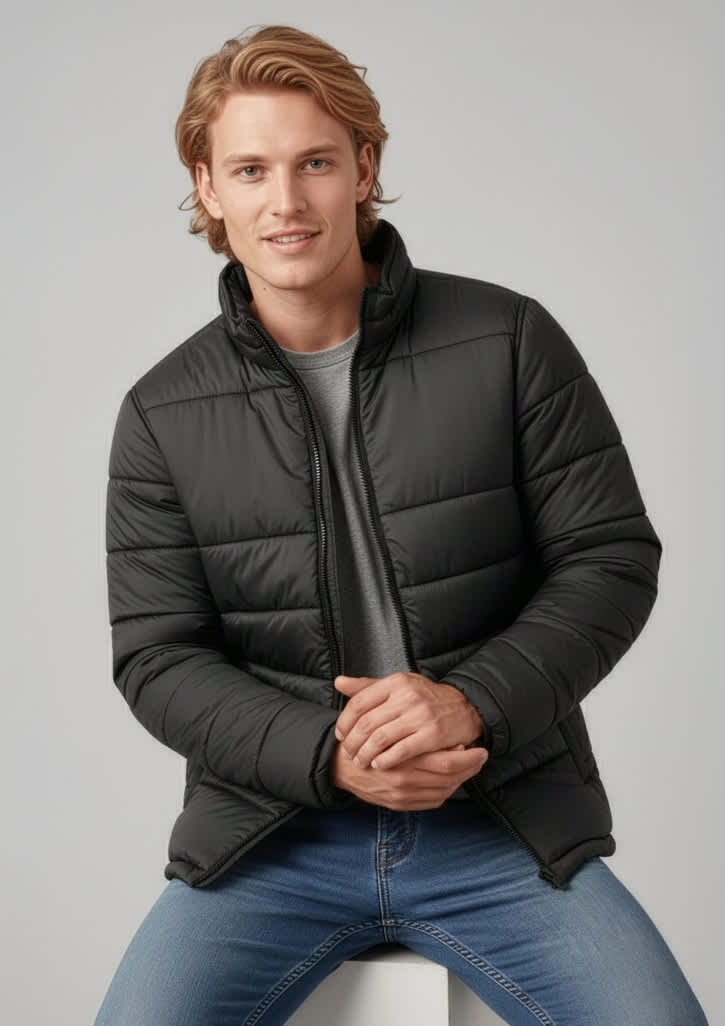 A man wearing a black puffer jacket, sitting on a white surface, with light jeans and a grey t-shirt. Mens Alpine Jacket - Image 1