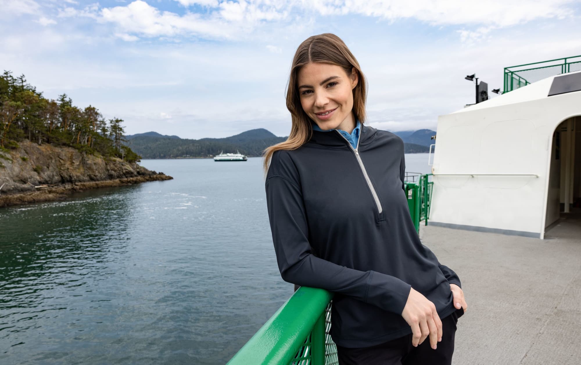 A half-zip hoodie in navy blue worn by a woman, featuring long sleeves and a subtle logo, against a scenic waterfront background. Ladies Daybreak Half Zip Hoodie - Image 1