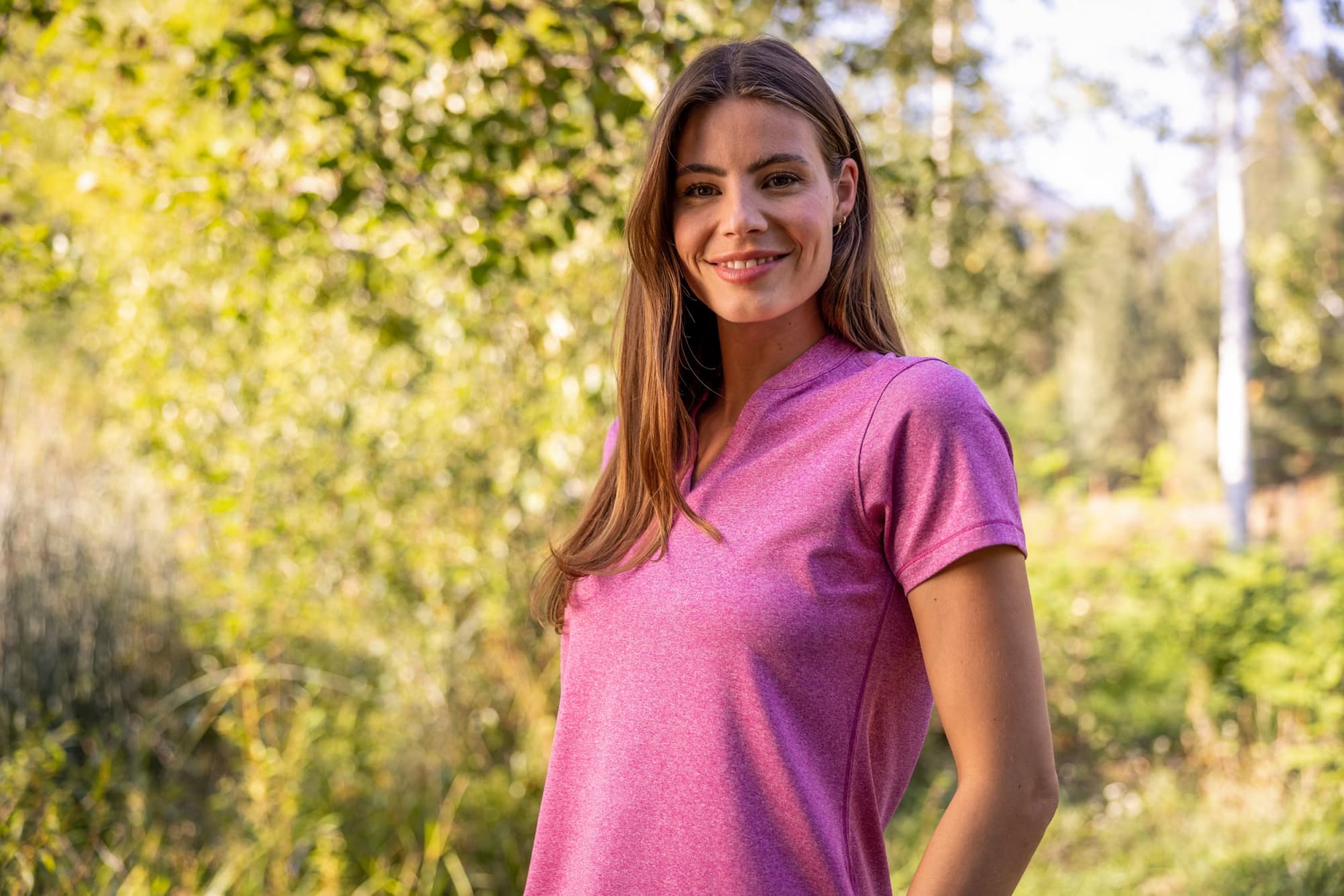 A woman is wearing a pink polo shirt in a natural outdoor setting with trees in the background. Ladies Forge Eco Heather Blade Polo - Image 1