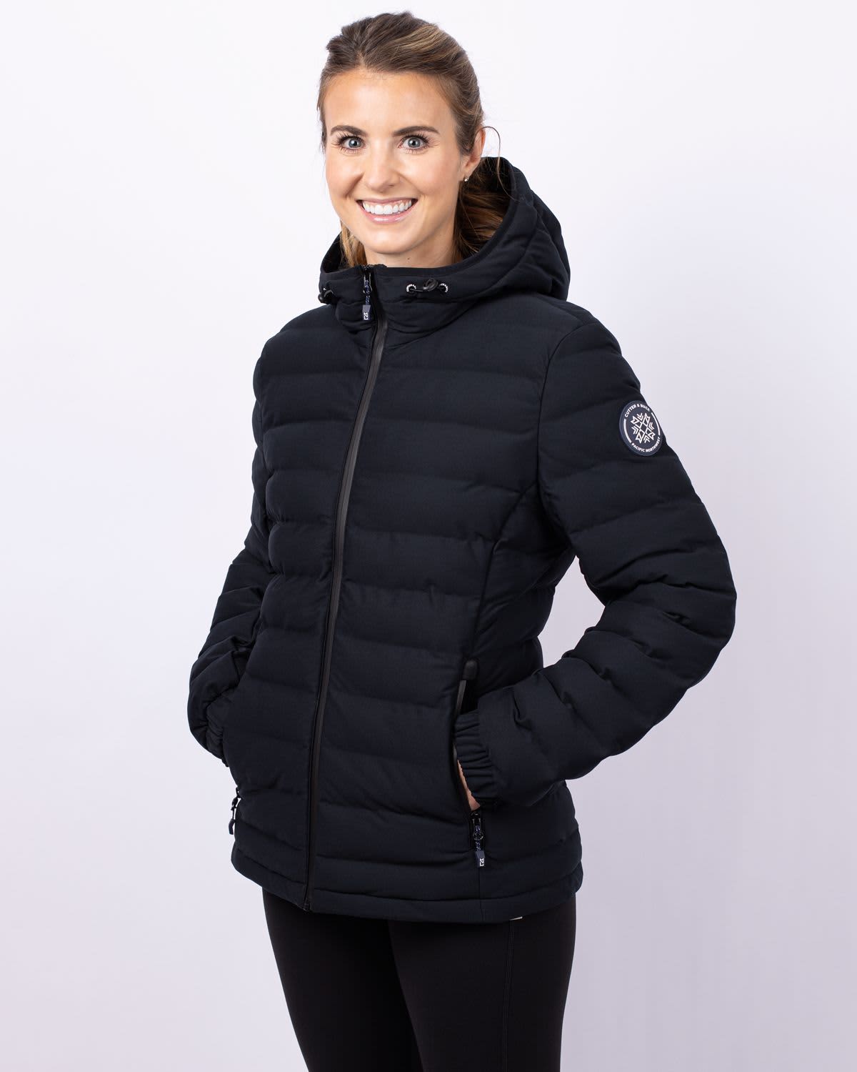 A black puffer jacket with a hood, featuring a logo on one sleeve, worn by a smiling woman. Ladies Mission Ridge Puffer Jacket - Image 1