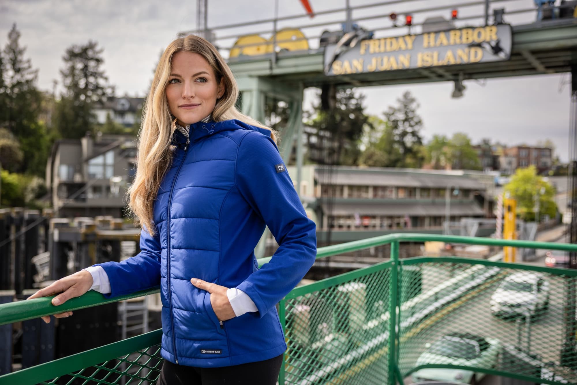 A woman wearing a blue soft shell jacket stands casually on a green railing with a scenic background. Ladies Evoke Eco Hybrid Jacket - Image 1