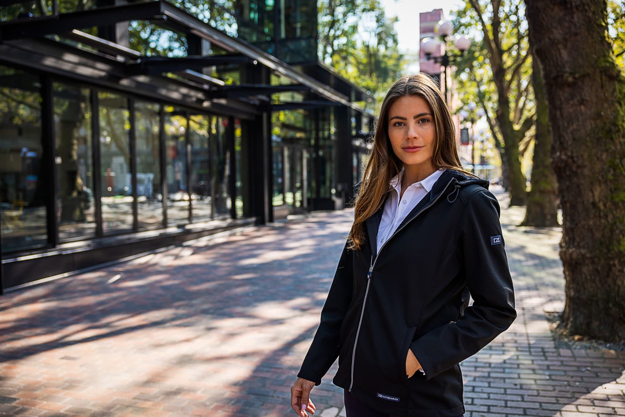 A woman stands outside wearing a black softshell jacket. The jacket features a hood and has a logo. Ladies Evoke Eco Softshell Jacket - Image 1
