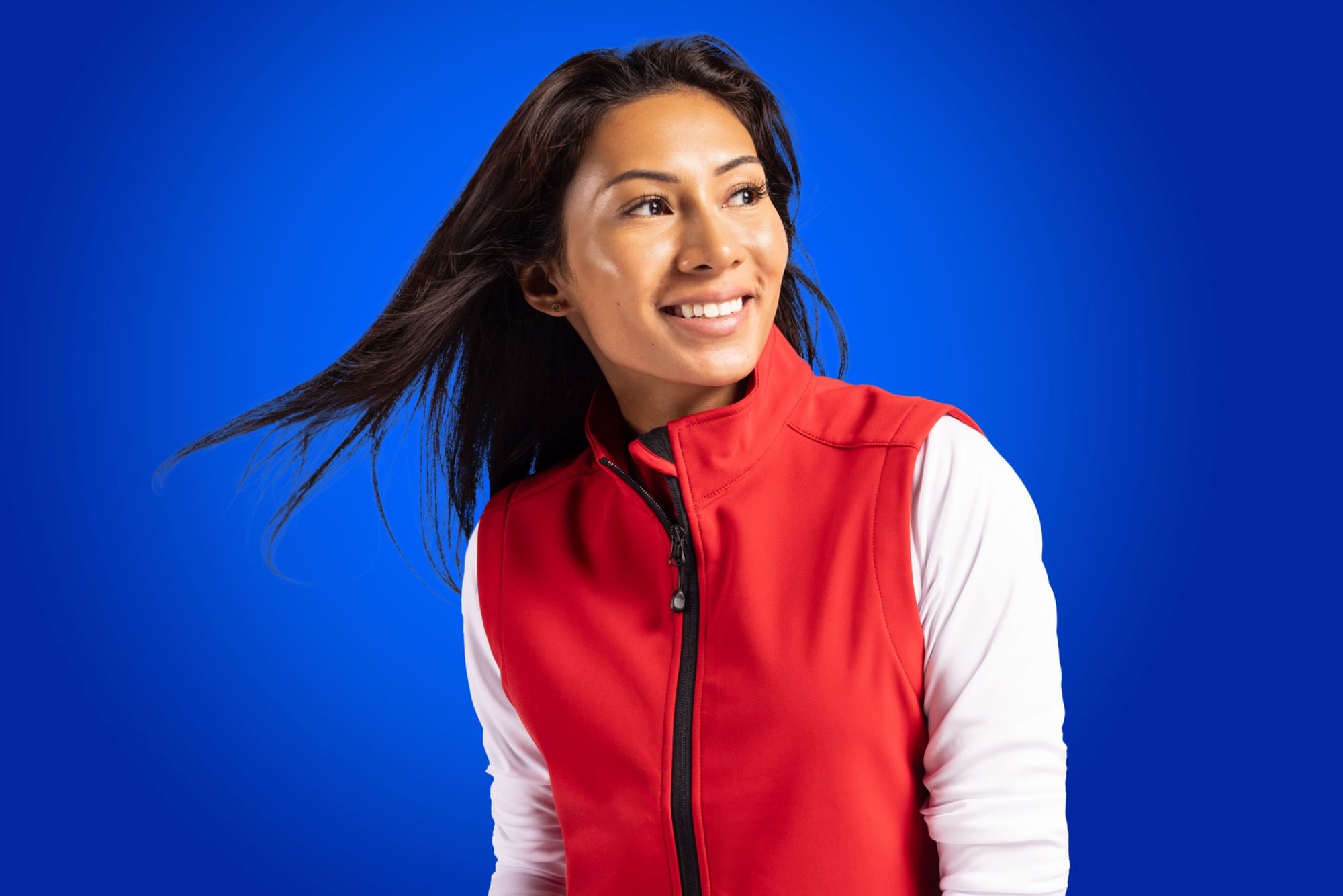 A woman wearing a red eco stretch vest with white sleeves, set against a blue background. The vest features a logo. Ladies Tempo Eco Stretch Vest - Image 1