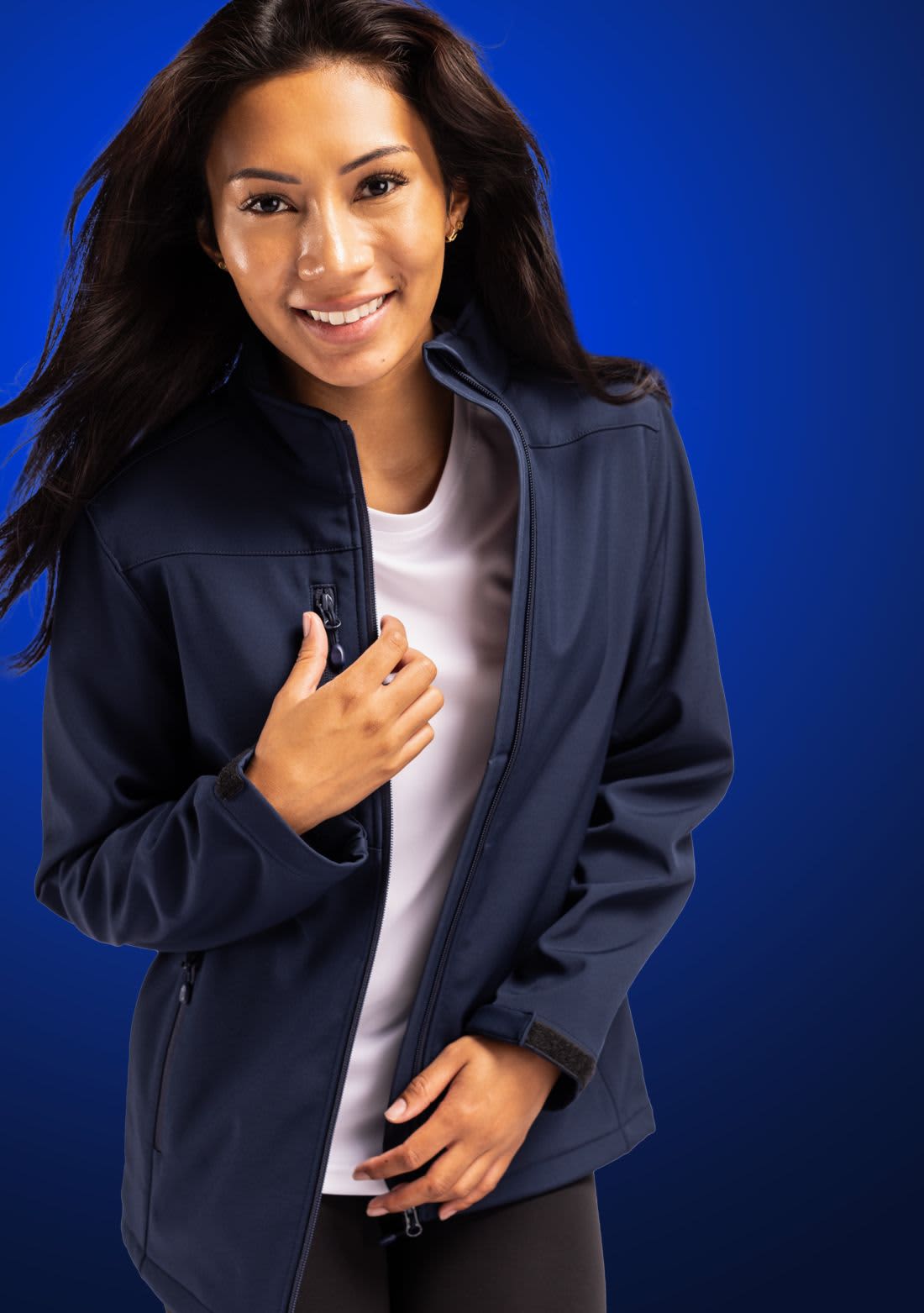 A woman wearing a navy blue softshell jacket with a zip and zippered pockets, smiling against a blue background. Ladies Tempest Eco Softshell Jacket - Image 1