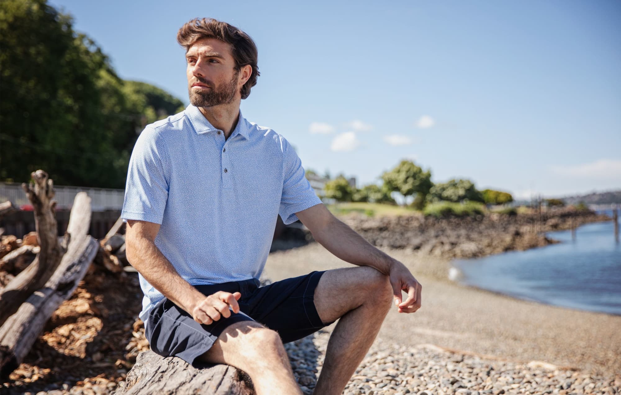 A man wearing a light blue botanical print polo shirt sits on a log by the water, set against a scenic background. Men's Virtue Eco Pique Botanical Print Polo - Image 1