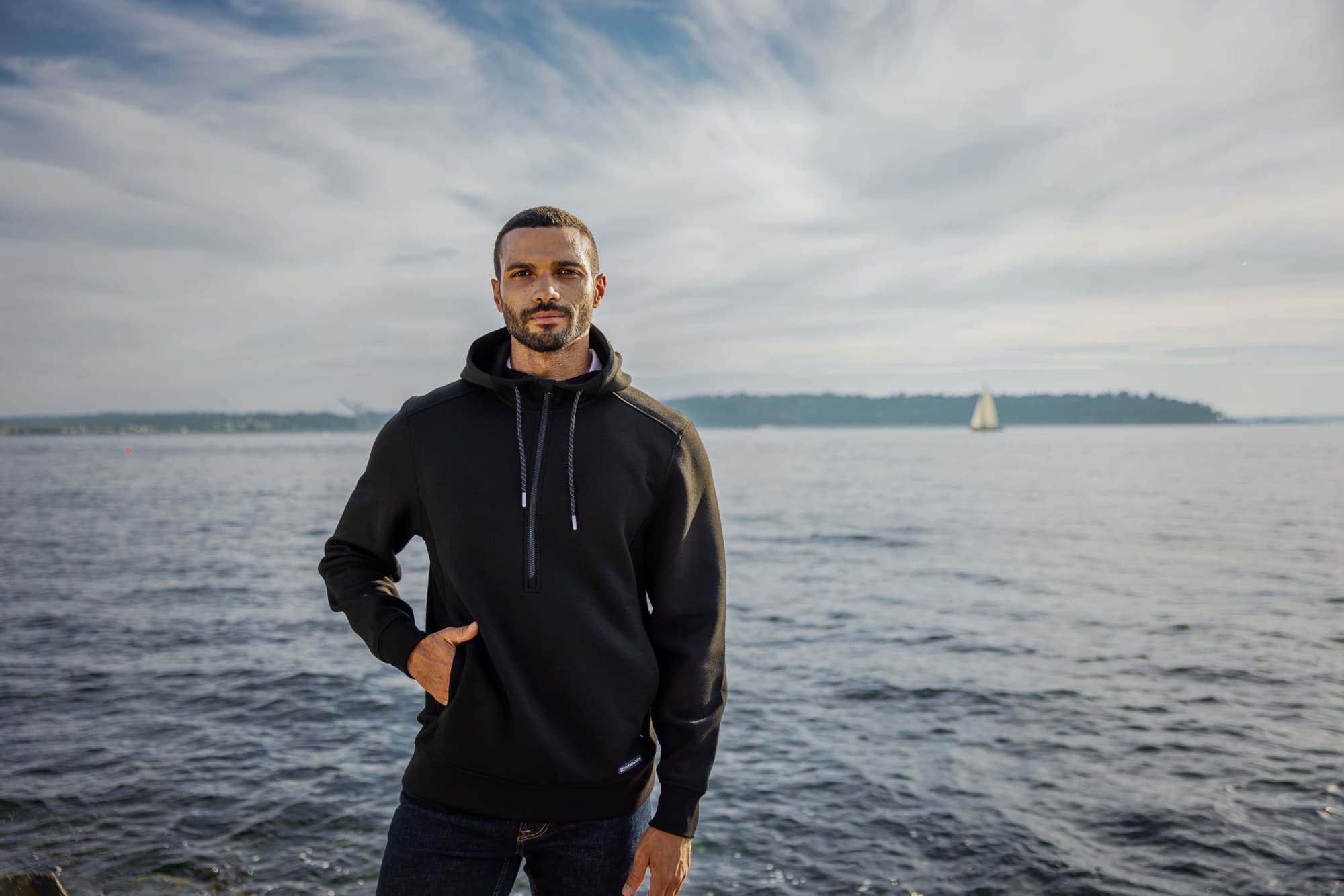 A man stands by the water wearing a black half zip hoodie, featuring a logo and a relaxed fit. Men’s Roam Eco Half Zip Hoodie - Image 1