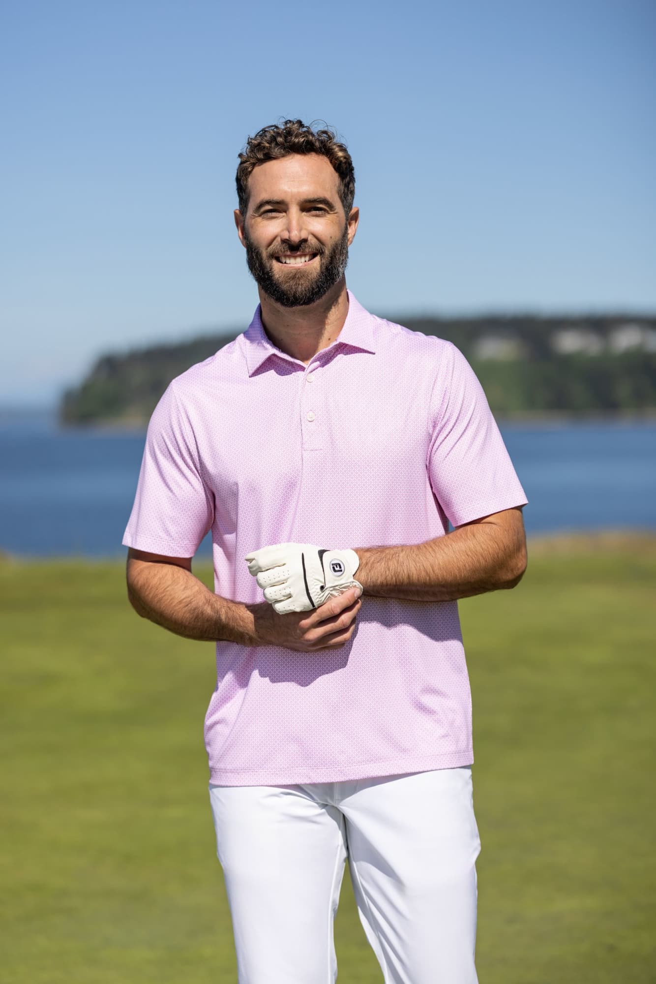 A man wearing a light pink polo shirt stands outdoors, with a golf glove on while posing against a scenic backdrop. Men’s Pike Eco Diamond Dot Polo - Image 1