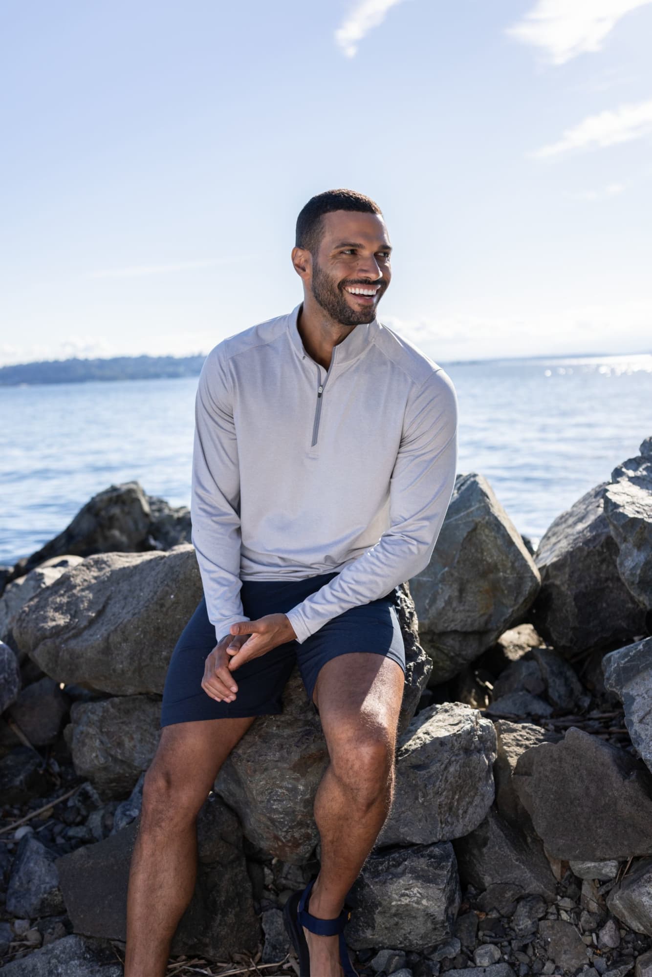 A man wearing a light grey quarter zip top sits on rocks by the water, smiling under a clear blue sky. Men's Coastline Quarter Zip - Image 1