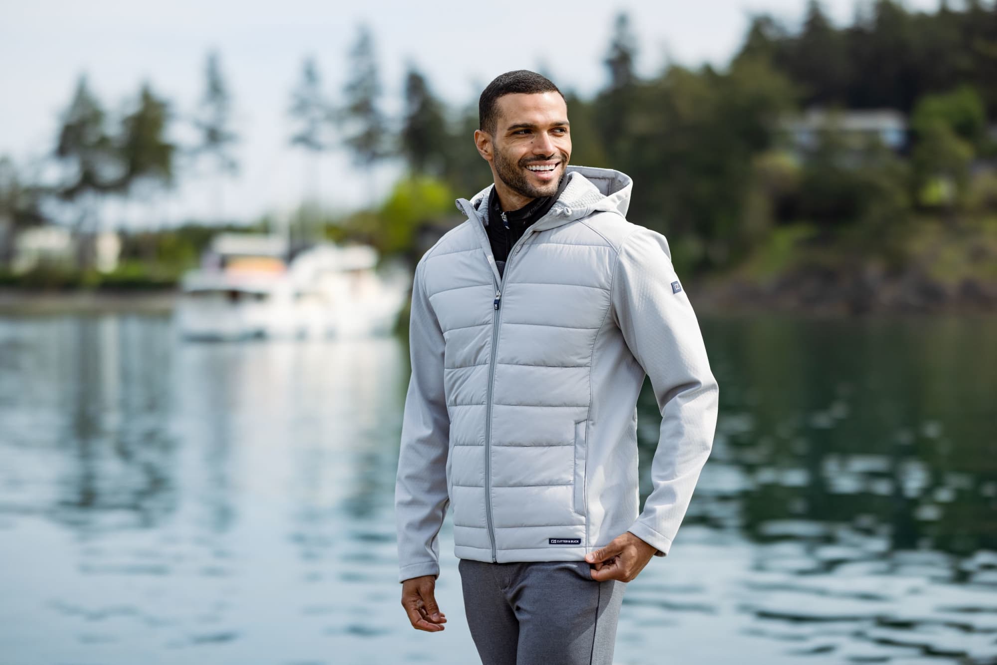 A man wearing a light grey hybrid jacket with a hood, smiling by the water, surrounded by trees and boats. Men’s Evoke Eco Hybrid Jacket - Image 1