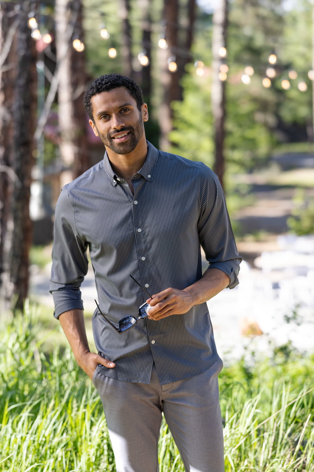 A man wearing a grey gingham shirt stands outdoors among greenery, holding sunglasses, with trees in the background. Men's Skyline Gingham Shirt - Image 1