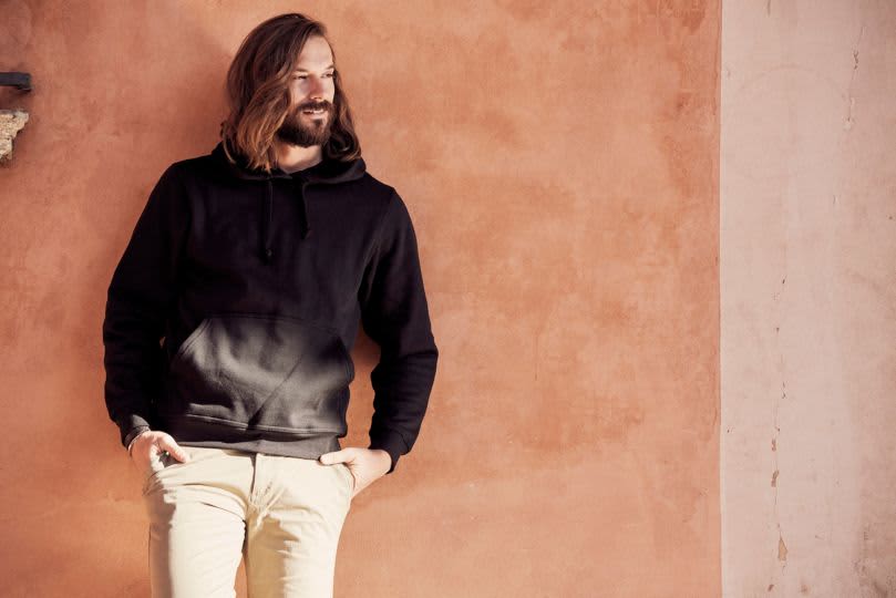 A black Stockholm hoodie displayed on a model against a warm-toned wall, featuring a front pocket and drawstring hood. Stockholm Hoodie - Image 1