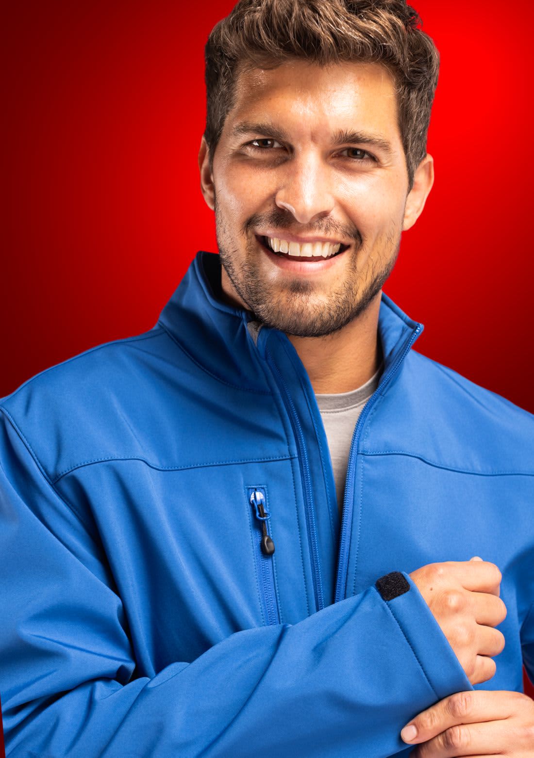 A man wearing a blue softshell jacket, smiling against a red background. The jacket has a logo on it. Men’s Tempest Eco Softshell Jacket - Image 1