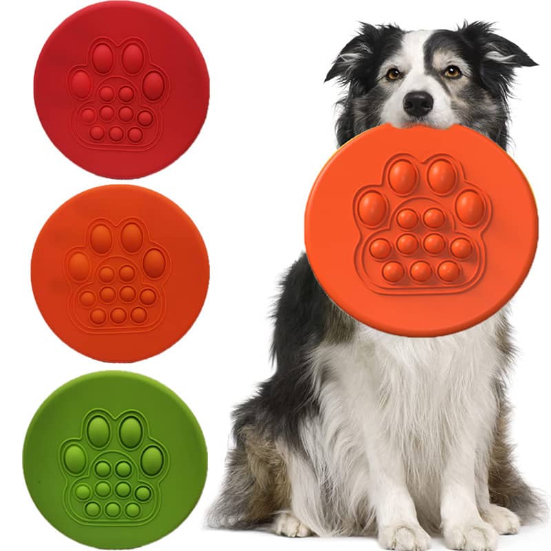 A dog holds a large orange flying disc, surrounded by red, orange, and green discs featuring a paw print design. Pet Flying Disc - Image 1