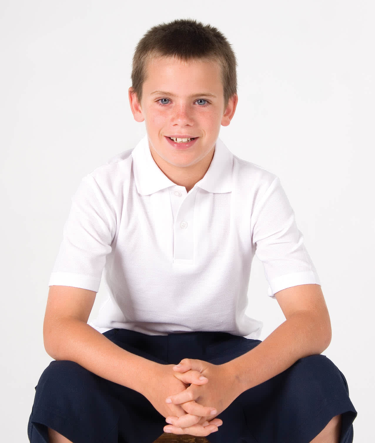 A white kid's pique polo shirt worn by a boy sitting, with dark shorts and a neutral background. The shirt has a logo. Kid's Pique Polo - Image 1