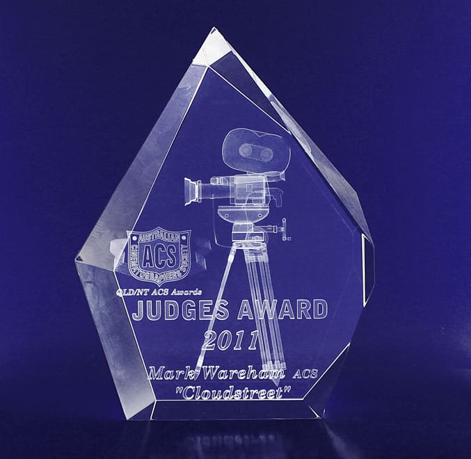 A clear, crystal trophy with a geometric design, featuring an engraved film camera and text, on a dark background. 3D Crystal Large Prestige (60mm deep) - Image 1
