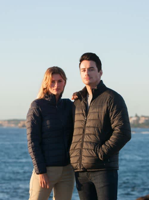 A couple stands by the water wearing navy puffer jackets. The scene is coastal with a clear sky. Puffer Jacket - Ladies - Image 1